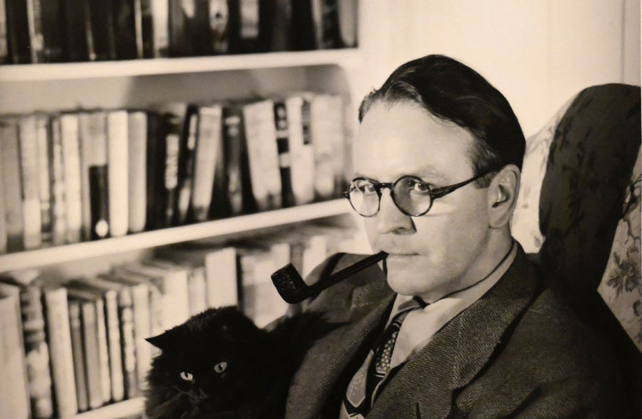 Raymond Chandler with his cat Taki sitting in a chair in his library with a pipe in his mouth